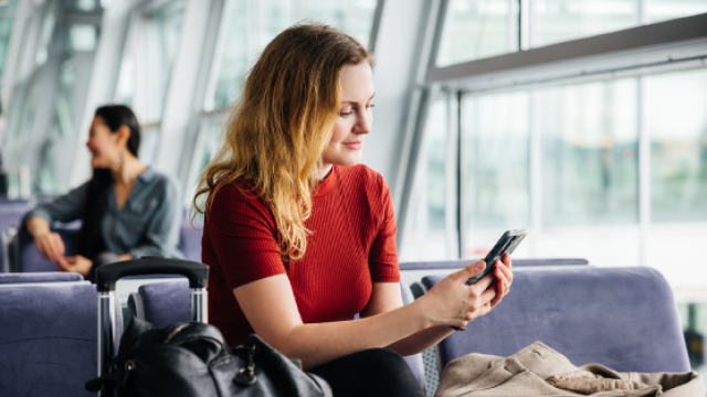 Young woman in red shirt waits at airport lounge seat checking her phone beside suitcase