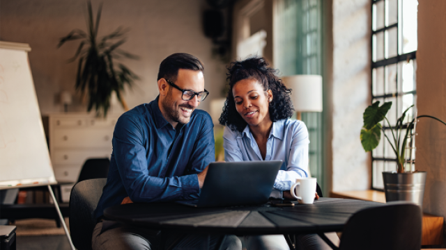 Two colleagues smiling over a laptop at a table in a sunlit office with plants
