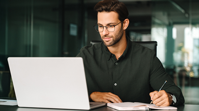 Photo of a man writing notes beside an open laptop in a modern office setting.
