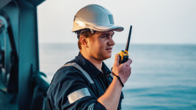 Ship officer in white hard hat and coveralls speaking into a handheld radio on deck
