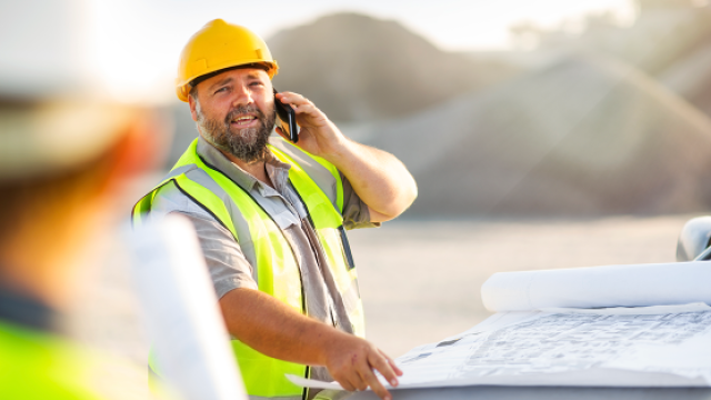 Smiling bearded construction worker in hard hat and vest checks blueprints and talks on phone