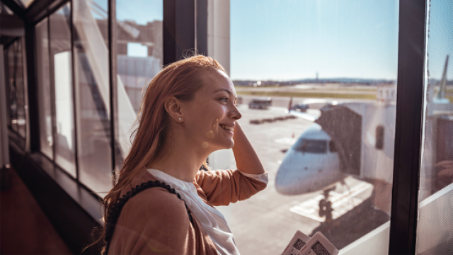 Young woman in airport terminal looking out at waiting airplane on the runway