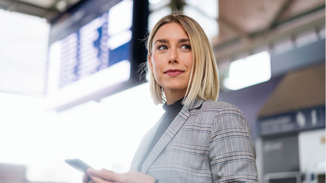 Photo of a blonde woman in a gray plaid blazer holding a phone at an airport terminal.