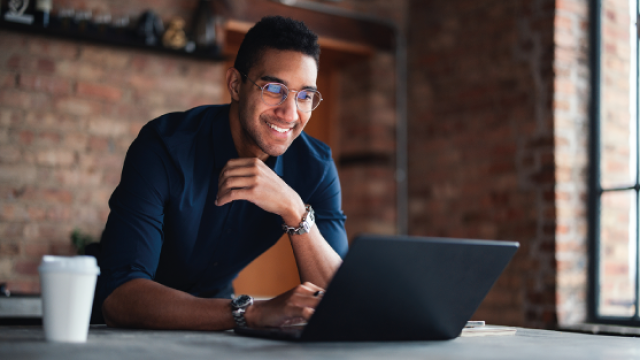 Smiling man in glasses working on laptop at brick-walled café table with coffee cup