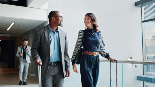 Photograph: Two coworkers walk a bright office hallway with laptops, one follows behind.