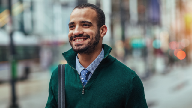 Photograph: smiling man in green sweater and tie on city sidewalk, blurred bus behind