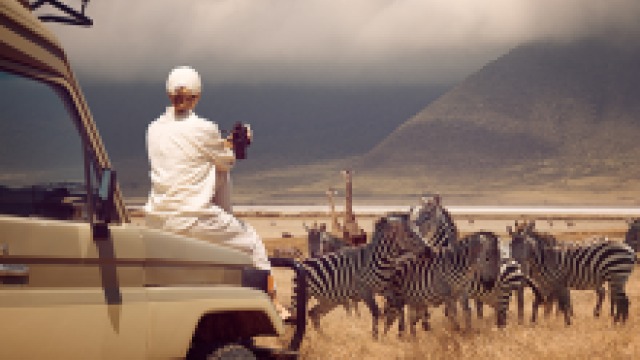 Man in safari truck photographing zebras on a grassy plain with distant mountains