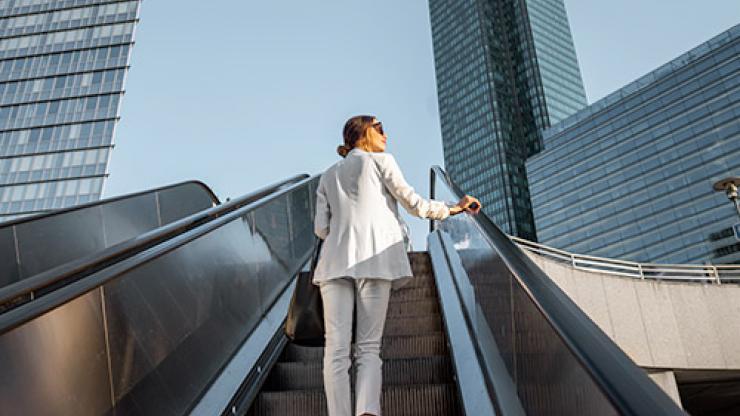 Stylish businesswoman in white suit going up on the escalator at the business centre outdoors with skyscrapers on the background
