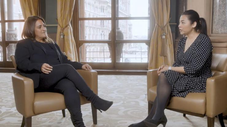 Two business women sit interview style in a hotel room