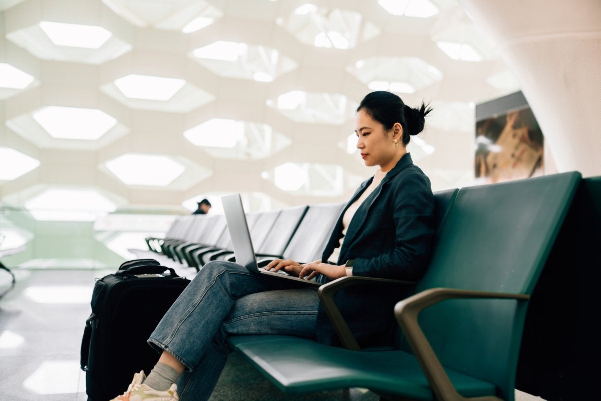 Profesional trabajando en una laptop desde la sala de espera de un aeropuerto mientras gestiona actividades relacionadas con viajes corporativos.