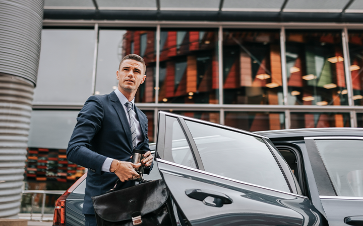 A man in a suit stands beside a car, holding a bag, in an urban setting with modern architecture.