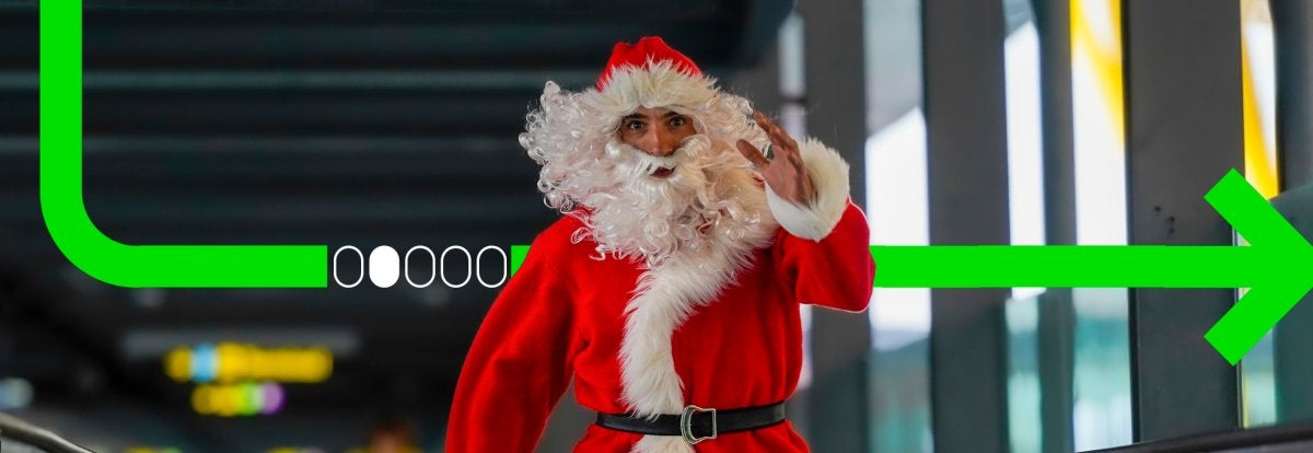 Persona disfrazada de Santa Claus caminando apresurado en un aeropuerto moderno mientras arrastra una maleta verde, transmitiendo humor y caos navideño.