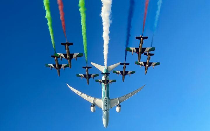 Etihad aircraft in flight with fighter jets expelling green, red and blue vapour