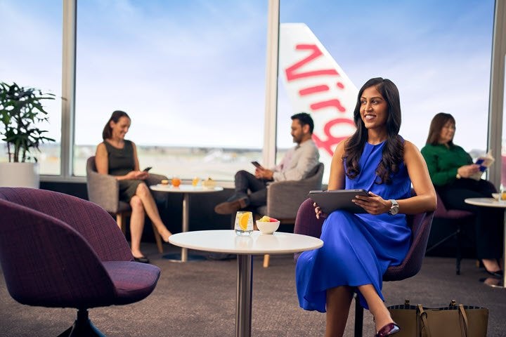 a young business woman in a blue silk dress sitting in the Virgin Australia lounge with an aircraft on the tarmac behind