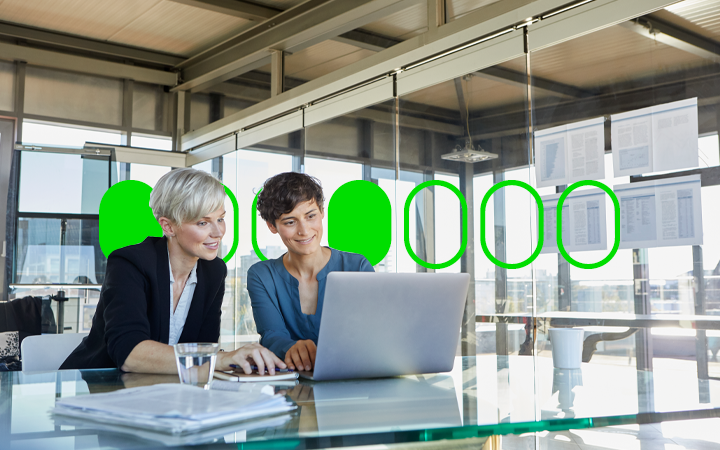Two ladies sitting at a desk in an office looking at a laptop screen together