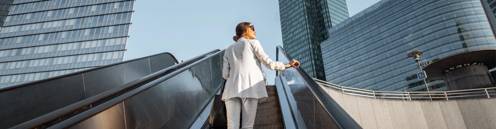 Businesswoman in white suit going up on the escalator at the business centre outdoors with skyscrapers on the background