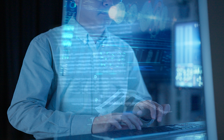A person in a light blue shirt typing on a laptop with digital data displayed on the screen.