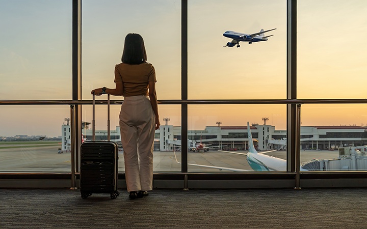 A woman with a suitcase gazes at an airplane taking off during sunset at an airport.