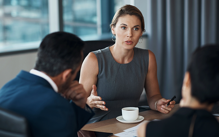 A woman in a gray dress speaks passionately with two colleagues in a meeting room.