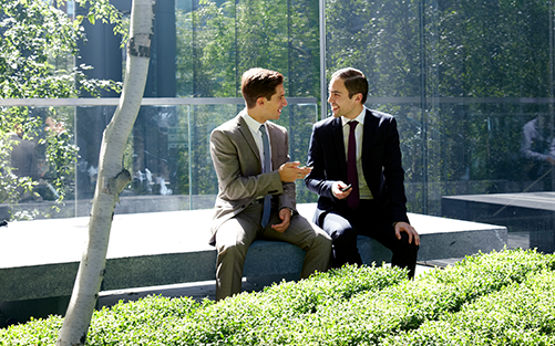 Two men in suits sitting on a stone bench outdoors, shaking hands in a green courtyard