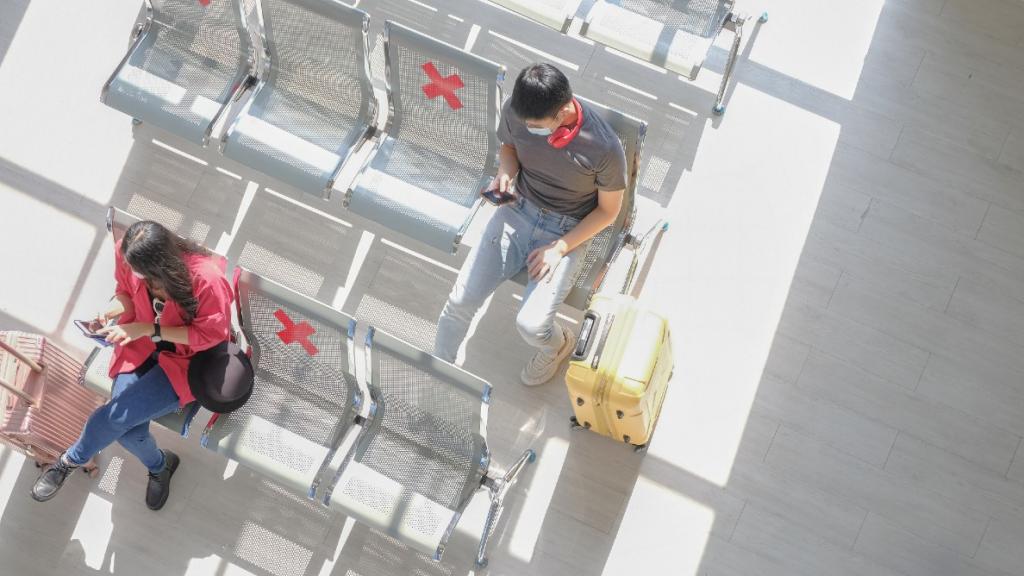 Birds eye view of people sitting in an airport with their suitcases, looking down at their phones