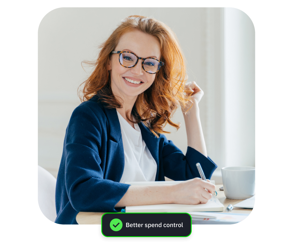 Photograph of a smiling redheaded woman with glasses sitting at a desk with papers and coffee