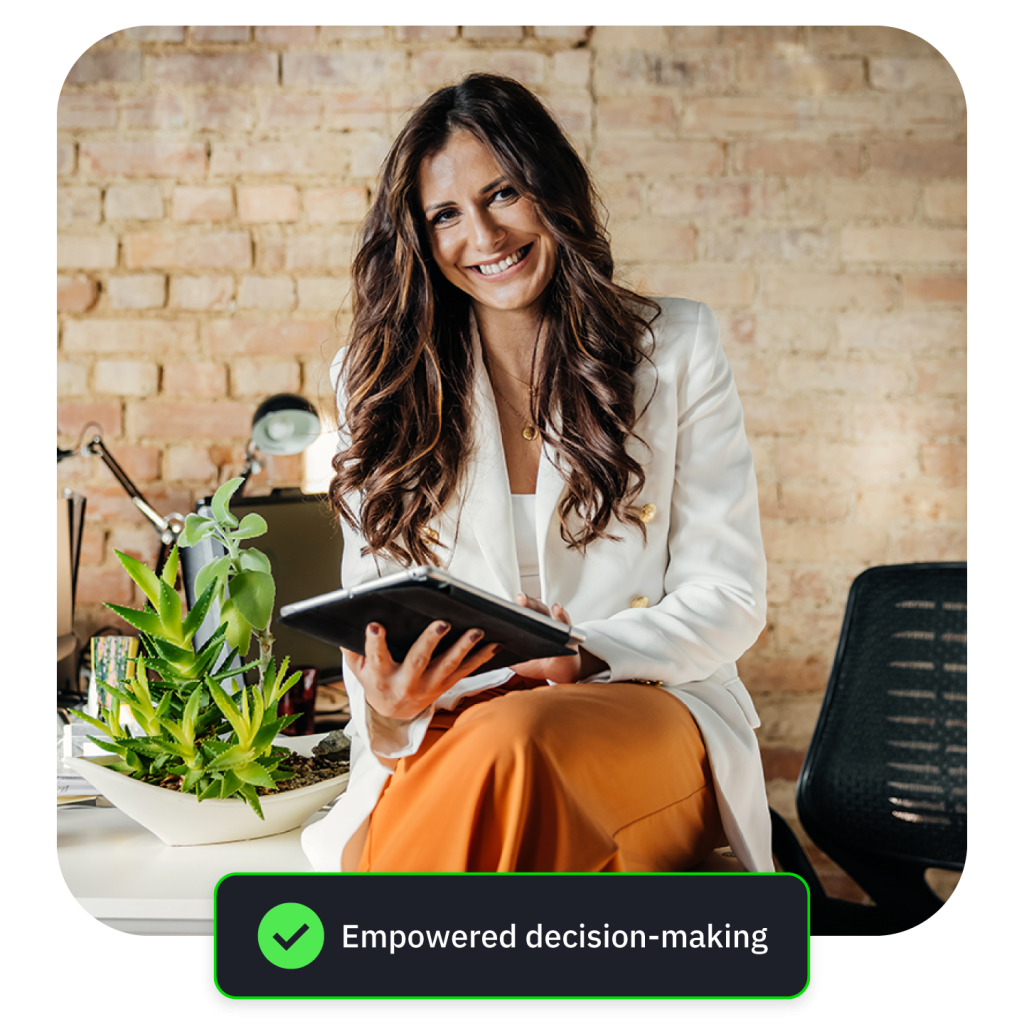 Smiling woman in white blazer and rust trousers holding a tablet, seated by a brick wall.