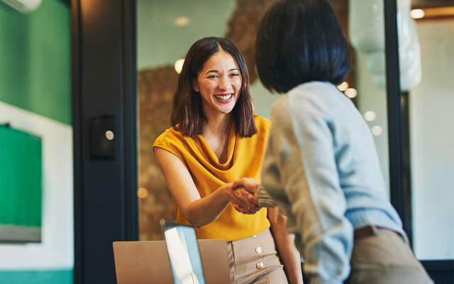Two professional women smiling and shaking hands across a desk in an office.