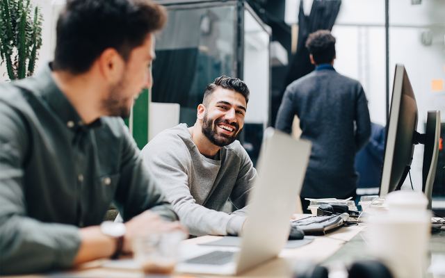 Two colleagues smiling at each other over laptops in a bright open office