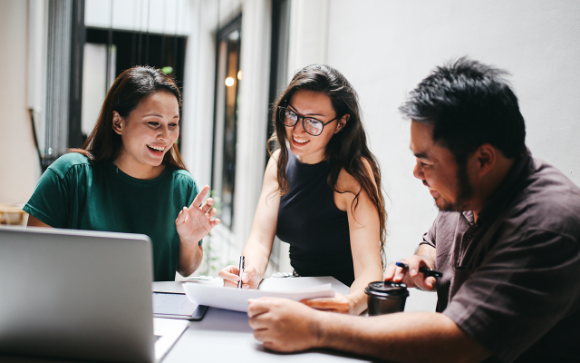 Three coworkers smiling and discussing papers by a laptop in an office.