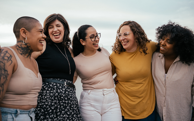 A group of five women smiling and laughing together outdoors in casual clothing.