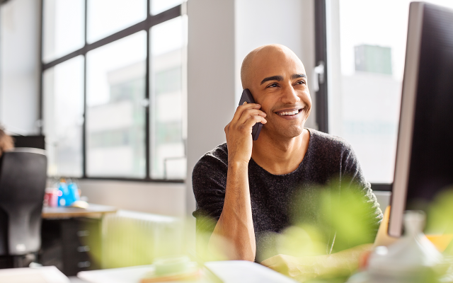 Smiling man at modern office desk talking on phone while looking at computer monitor