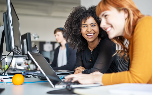 Photograph of two women smiling and collaborating on laptops in a bright modern office.