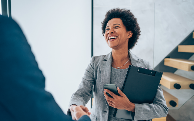 Smiling woman in gray blazer holds folder and shakes hands in modern office