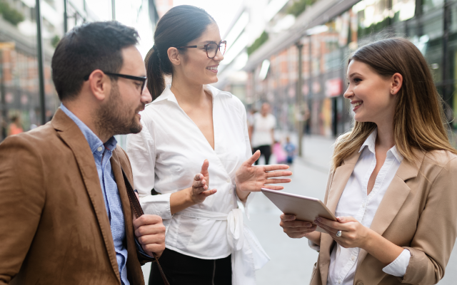 Three blurred businesspeople on city sidewalk, one gesturing, one with tablet, one giving thumbs up