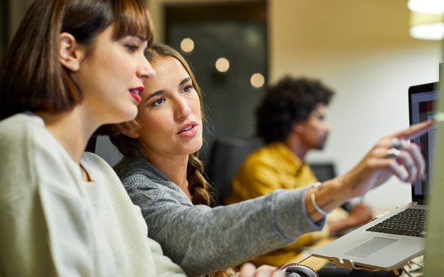 Two women in office collaborate over a laptop, one pointing toward the screen.