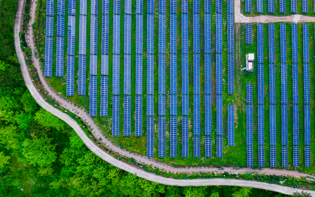 Drone photo of blue solar panel arrays along a winding dirt road beside green trees