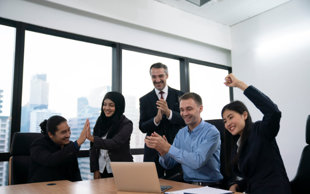 A diverse group of professionals celebrating success in a modern office with a laptop.