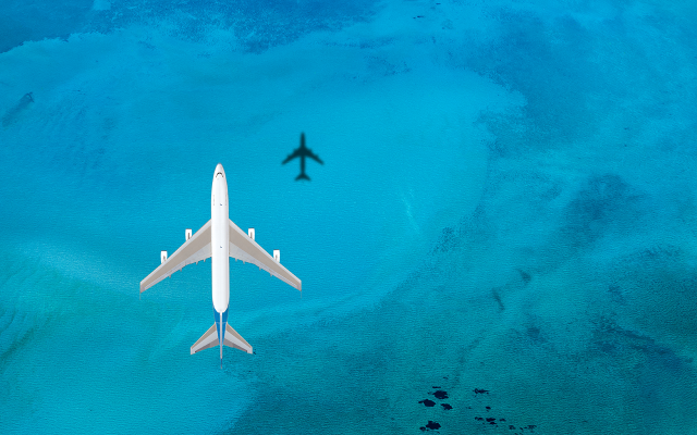 White passenger jet and its shadow soaring above clear turquoise ocean waters.