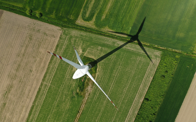 Aerial view of a white wind turbine casting a shadow over green and brown farmland