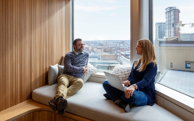 Two colleagues chatting on a cushioned window bench in modern office overlooking city skyline.