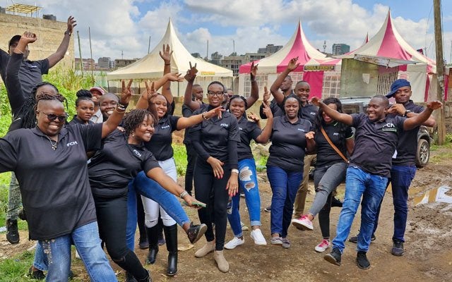 Group of adults in matching shirts smiling and raising fists in front of colorful festival tents