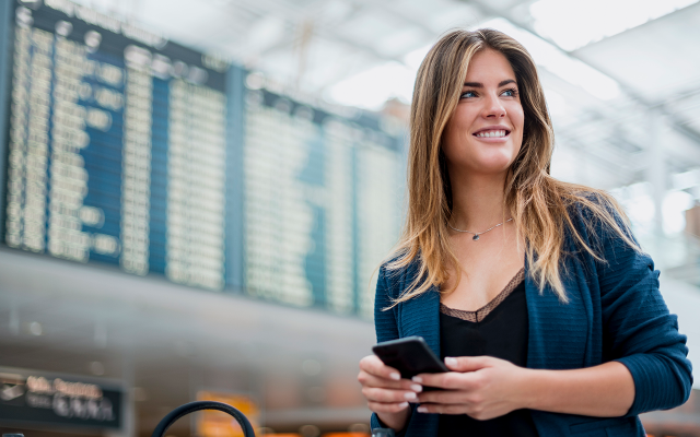 Smiling woman holding phone in airport terminal with departures board in background.