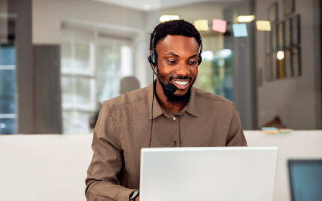 Young man wearing a headset, smiling while working on a laptop in a bright office setting