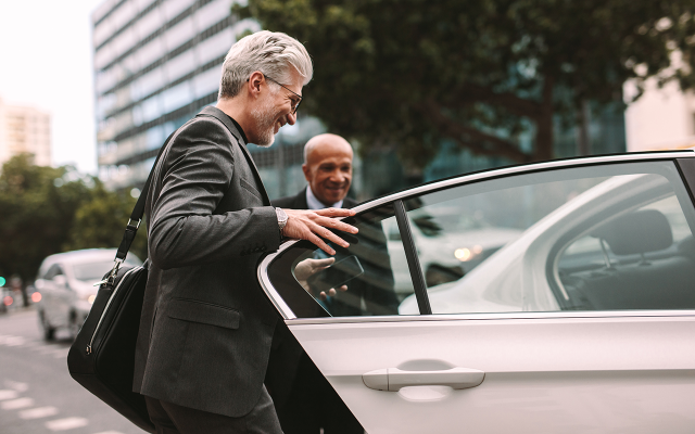 Younger chauffeur holding open car door as older businessman in suit enters