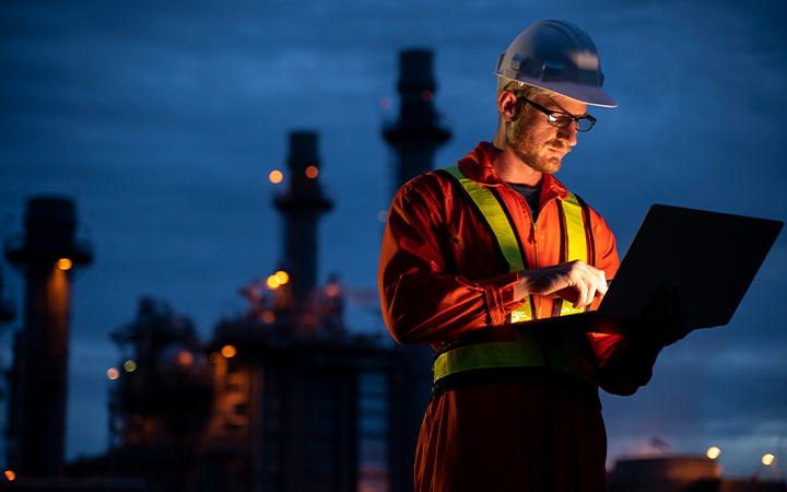 Man wearing hard hat looking at laptop standing in front of power station at night