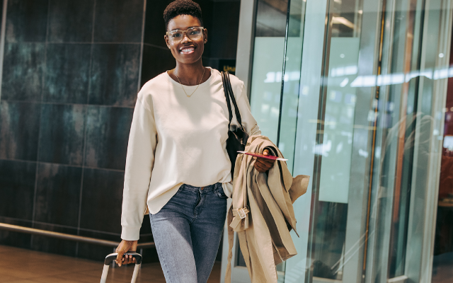 Woman in white blouse and jeans holding trench coat and purse in glass-walled corridor