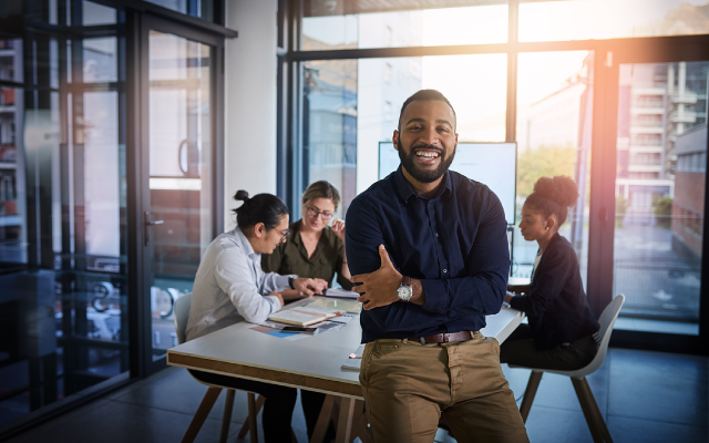Bearded man with arms crossed smiling in sunlit office, two colleagues collaborating over a laptop