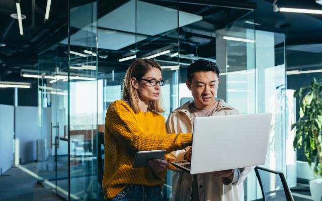 Photo of two coworkers working on a laptop in a glass-walled office.