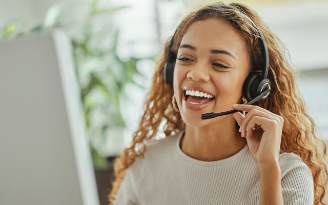 Young woman with curly hair wearing headset, smiling at laptop in bright office